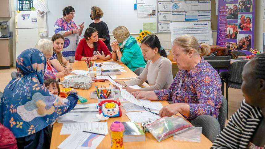 A group of eight woman from different ethnic backgrounds sit around a table talking to one another. Another two women stand in the background, having a conversation. 