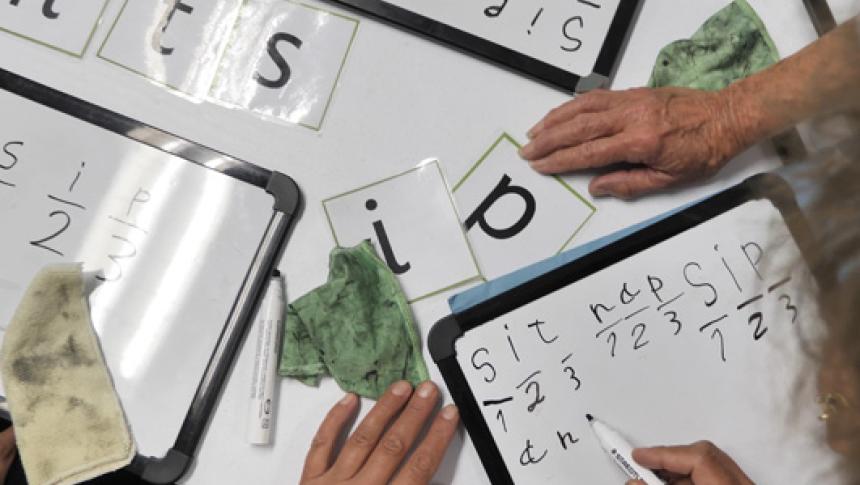 A table covered in letters and minature whiteboards. Two sets of hands can be seen practicing creating words on the whiteboards. 
