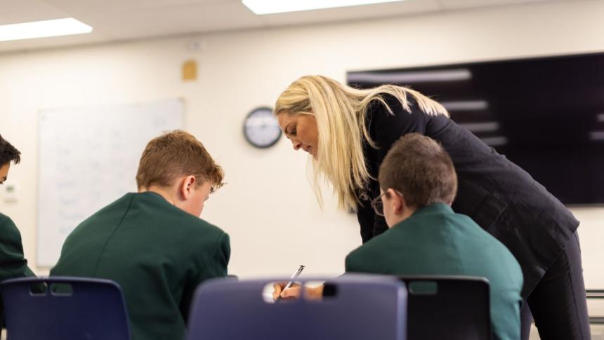 Teacher leaning over desk giving feedback to 2 secondary school students.