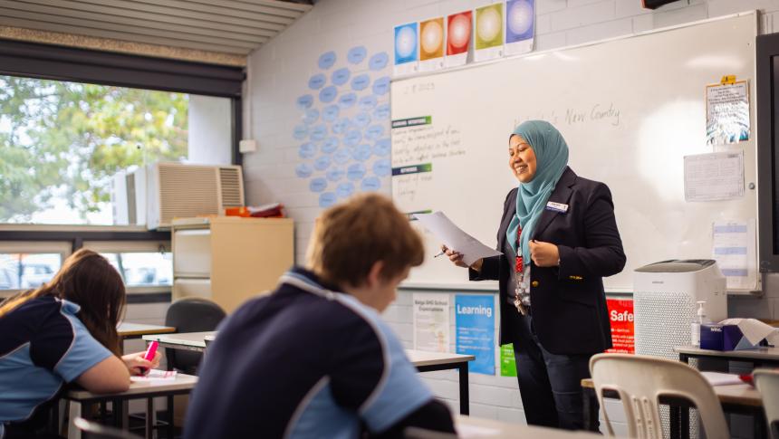 Teacher standing at the front of the classroom reading from a page