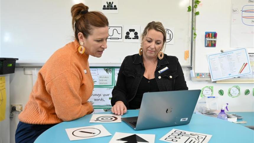 Two women look at a laptop, with black and white images scattered on a table in front of them.