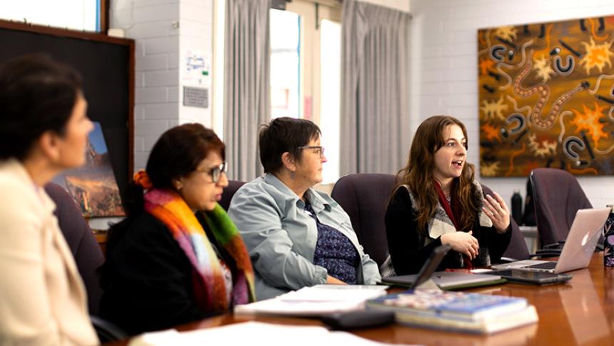 Four staff members sit at a boardroom table. One is speaking and the others are listening.