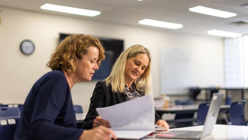 Two female teachers sit side by side at a desk, they are looking at pages in a notebook, and an open laptop is in front of them